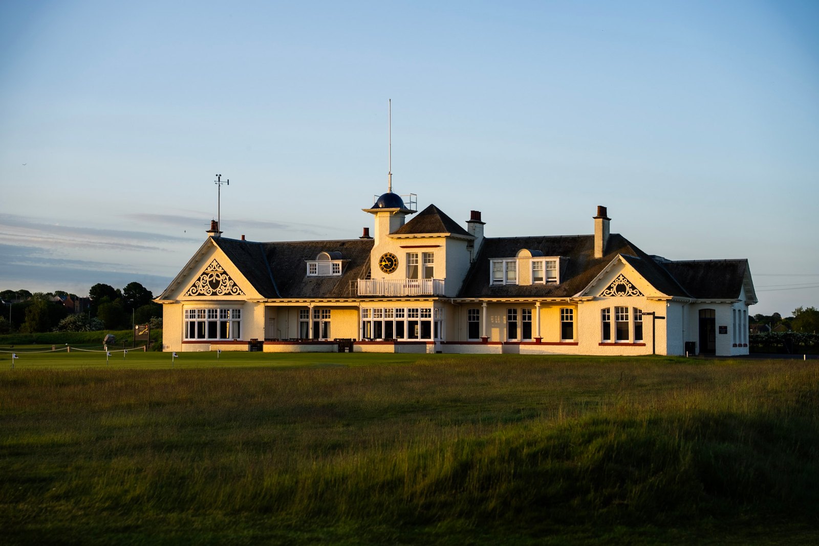 The Panmure clubhouse full frontage in low evening sun, the clock tower and gables lit gold, seen across the closing green