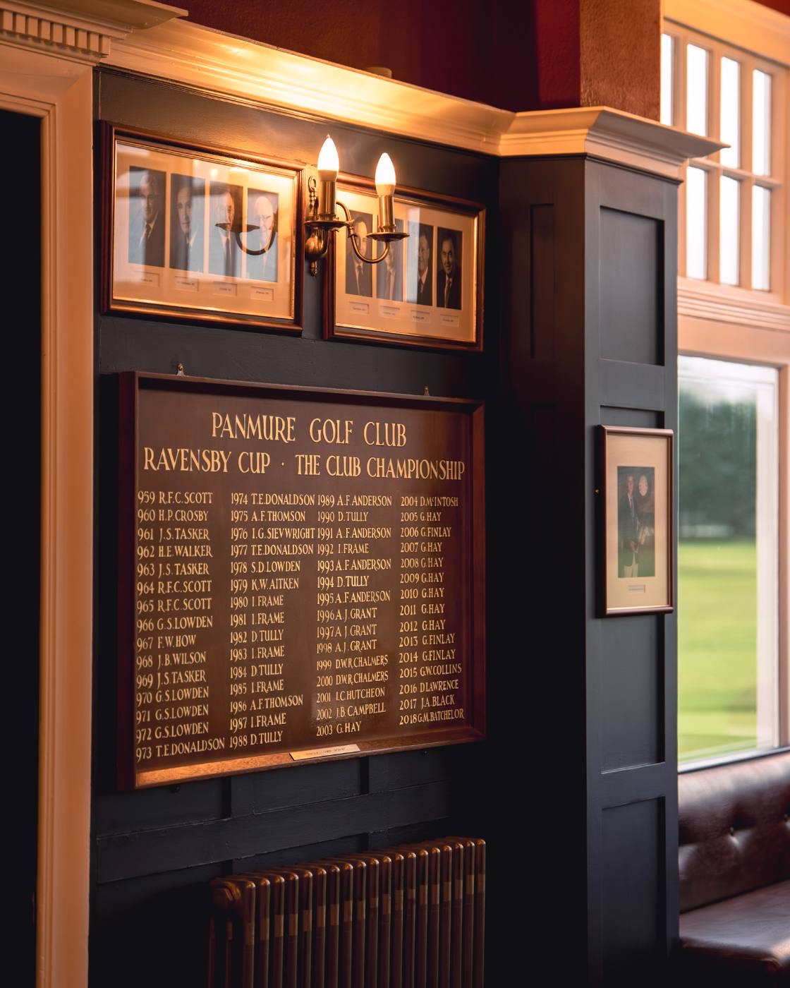 The Ravensby Cup honours board in the clubhouse, gilt lettering on dark panelling under a pair of wall lights, with portraits of past captains above