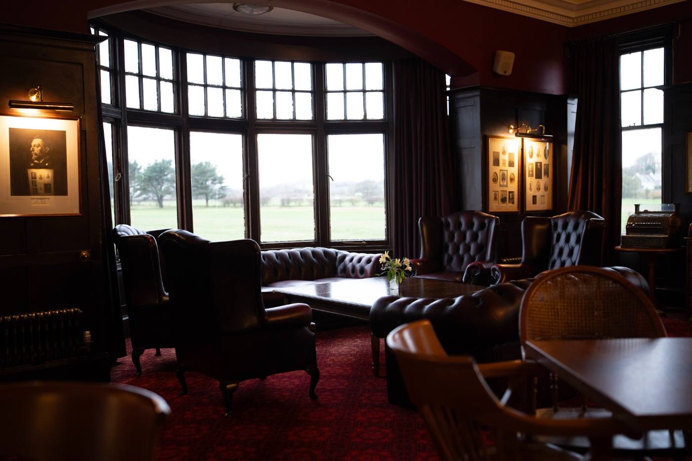 A bay-window lounge with deep leather chesterfields, a framed portrait and a lit display cabinet