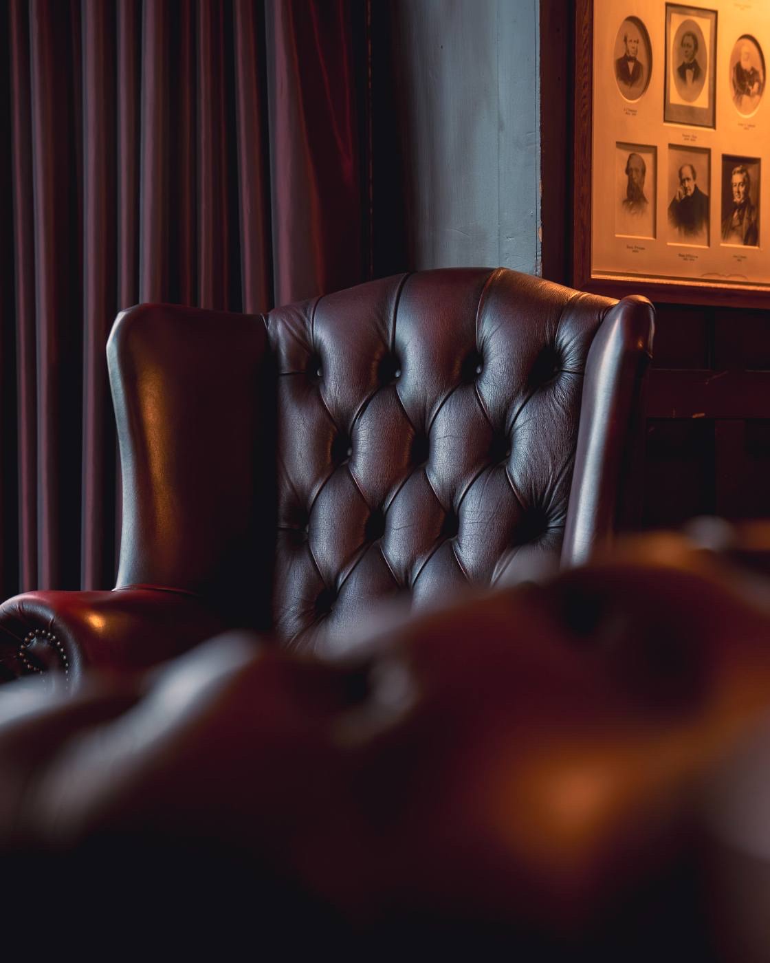 A deep-buttoned leather wing armchair in a panelled lounge, framed sepia portraits behind