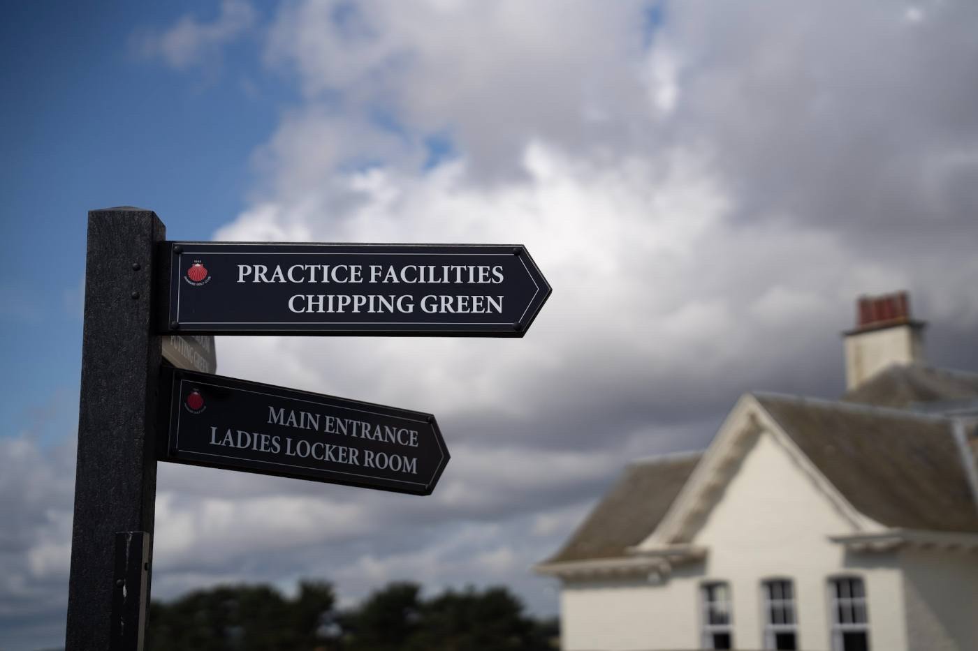 A fingerpost with arms reading Practice Facilities, Chipping Green, Main Entrance and Ladies Locker Room, with the clubhouse behind